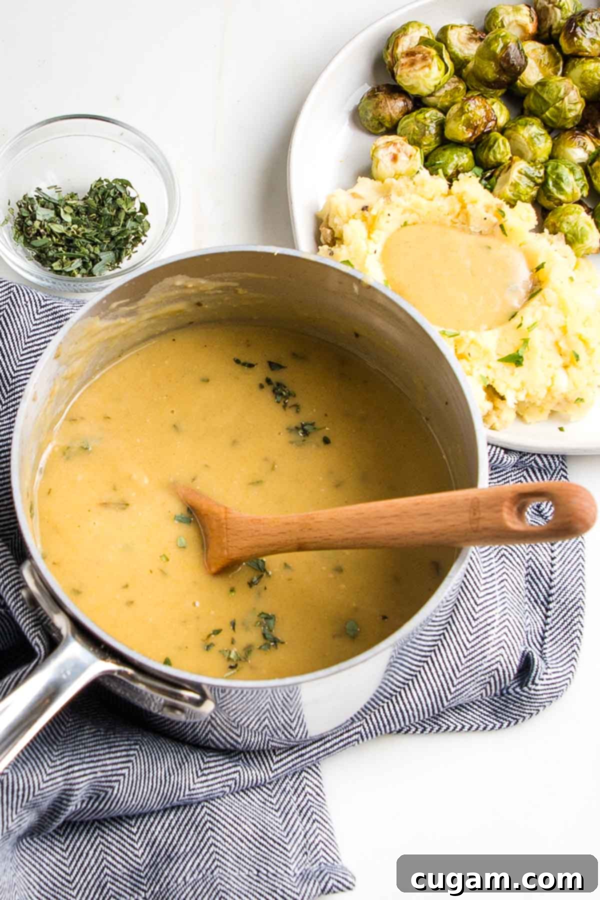 Small saucepan filled with golden gravy, a wooden spoon stirring, next to a plate of mashed potatoes and gravy with brussels sprouts.
