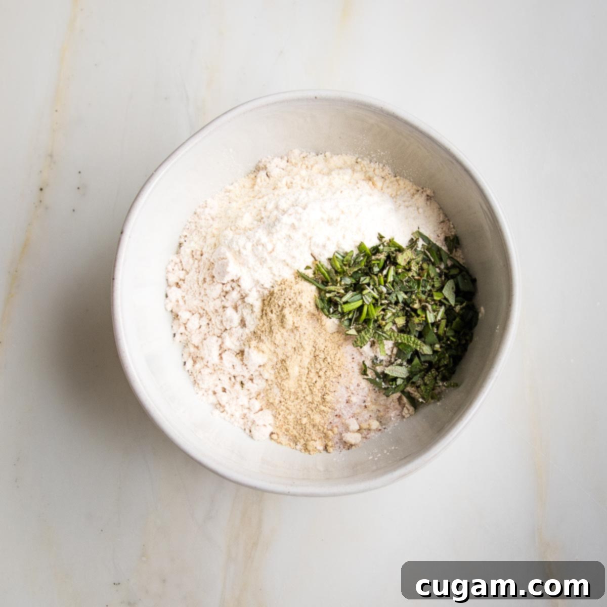 Small white bowl with flour, fresh herbs, arrowroot powder, salt, white pepper, garlic powder and onions powder.