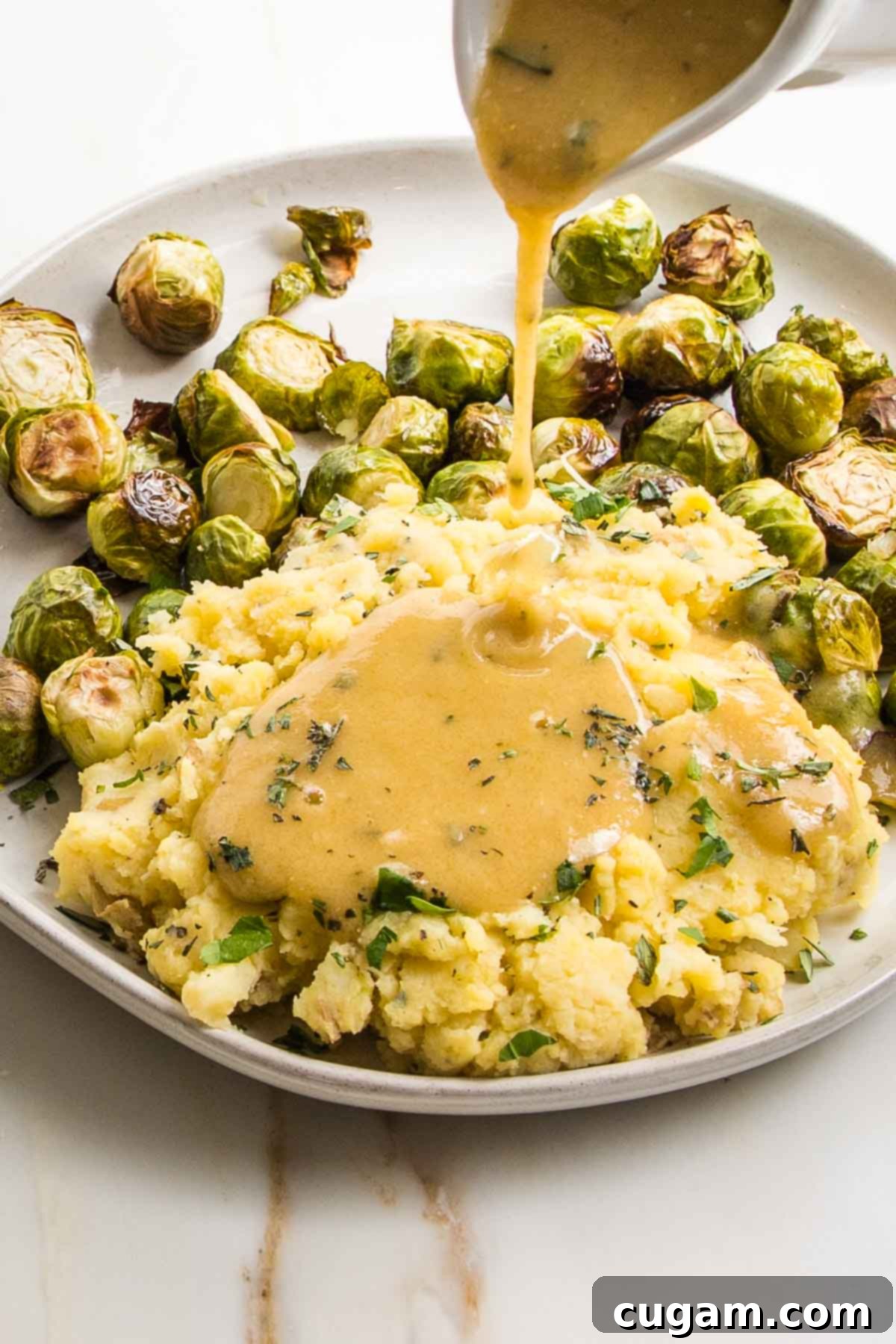 White pitcher pouring vegan gravy over a plate of mashed potatoes and brussels sprouts