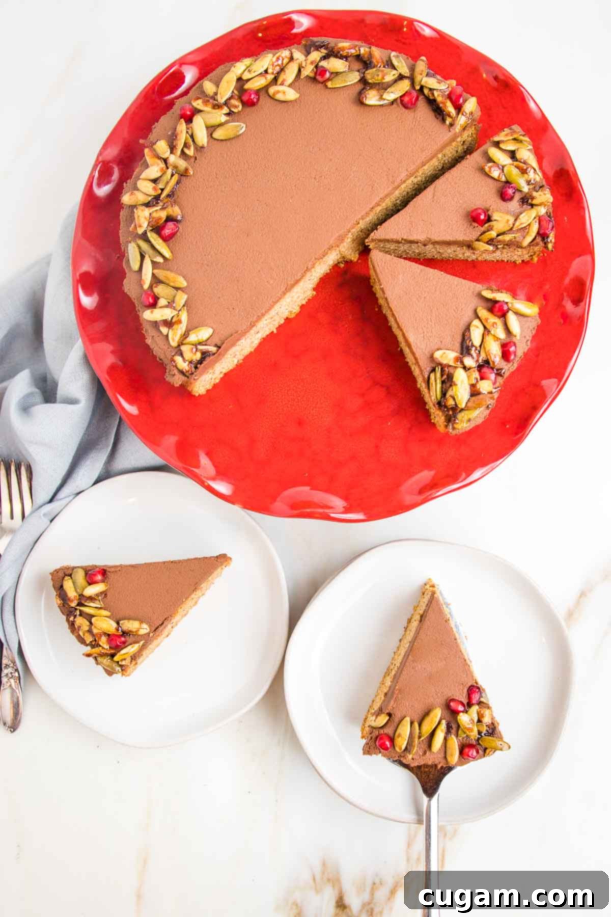 Pumpkin Cake with rich chocolate frosting, elegantly decorated with vibrant candied pepitas and ruby-red pomegranate seeds. The cake sits proudly on a red pedestal, with two inviting slices on white plates awaiting eager tasters in the foreground.