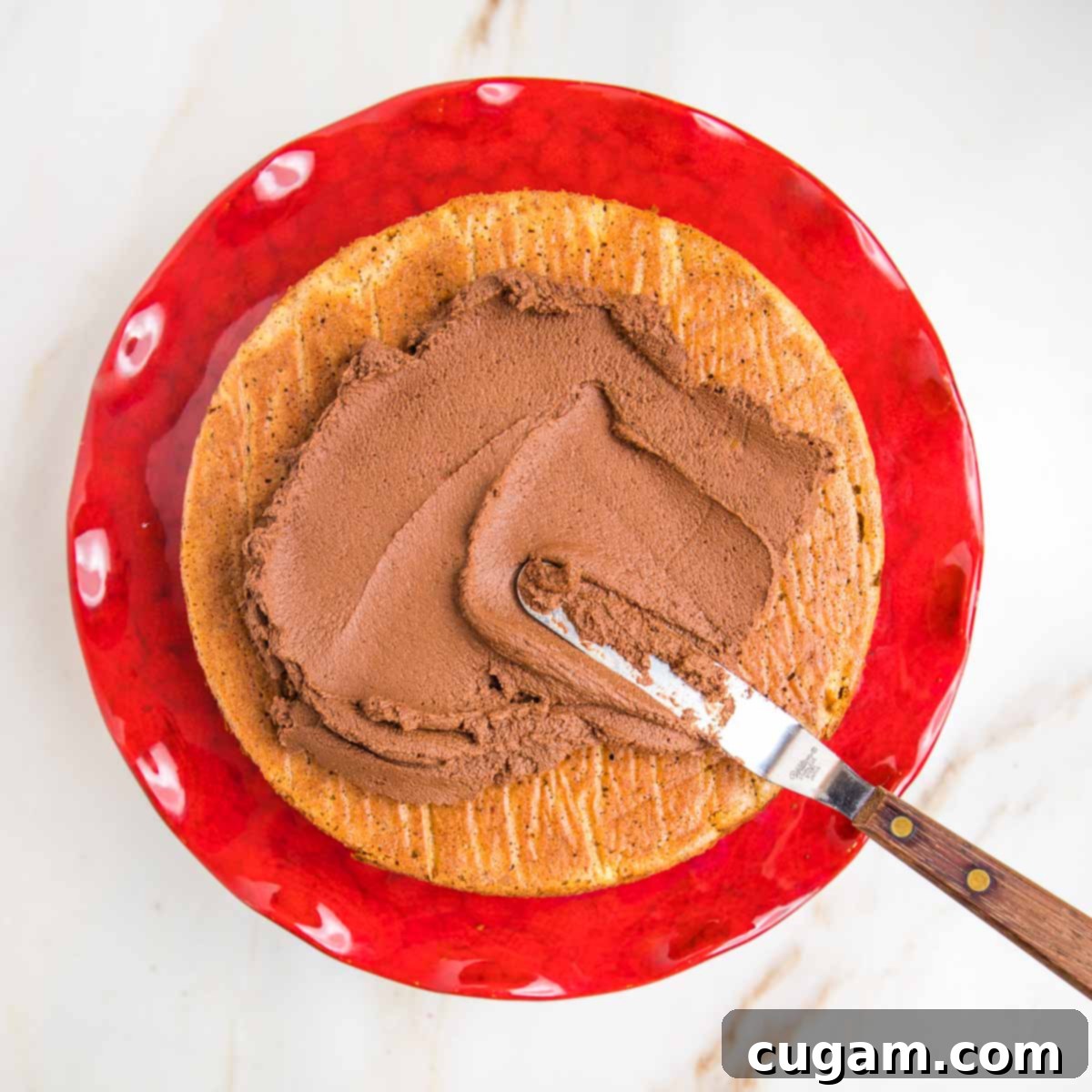A pumpkin cake set on a vibrant red pedestal, with an offset spatula gracefully spreading rich chocolate frosting across its top surface.