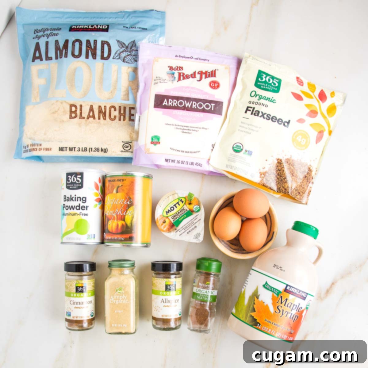 A collection of pumpkin cake ingredients laid out in their packaging or small bowls: almond flour, arrowroot powder, ground flax, baking powder, a can of pumpkin puree, a small container of applesauce, a bowl of three eggs, maple syrup, and four jars of various spices including ginger, allspice, cinnamon, and nutmeg.