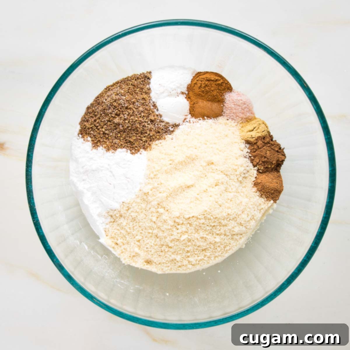 A clear glass bowl holding almond flour, arrowroot powder, ground flax, and small scoops of various brown pumpkin spices.