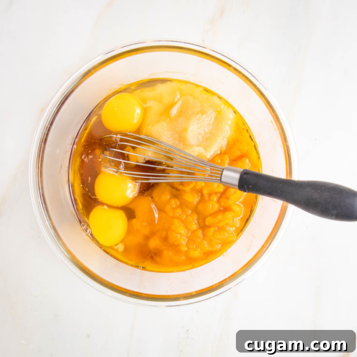 A glass bowl containing three eggs, with applesauce, maple syrup, and pumpkin puree nearby, along with a whisk, ready for mixing.
