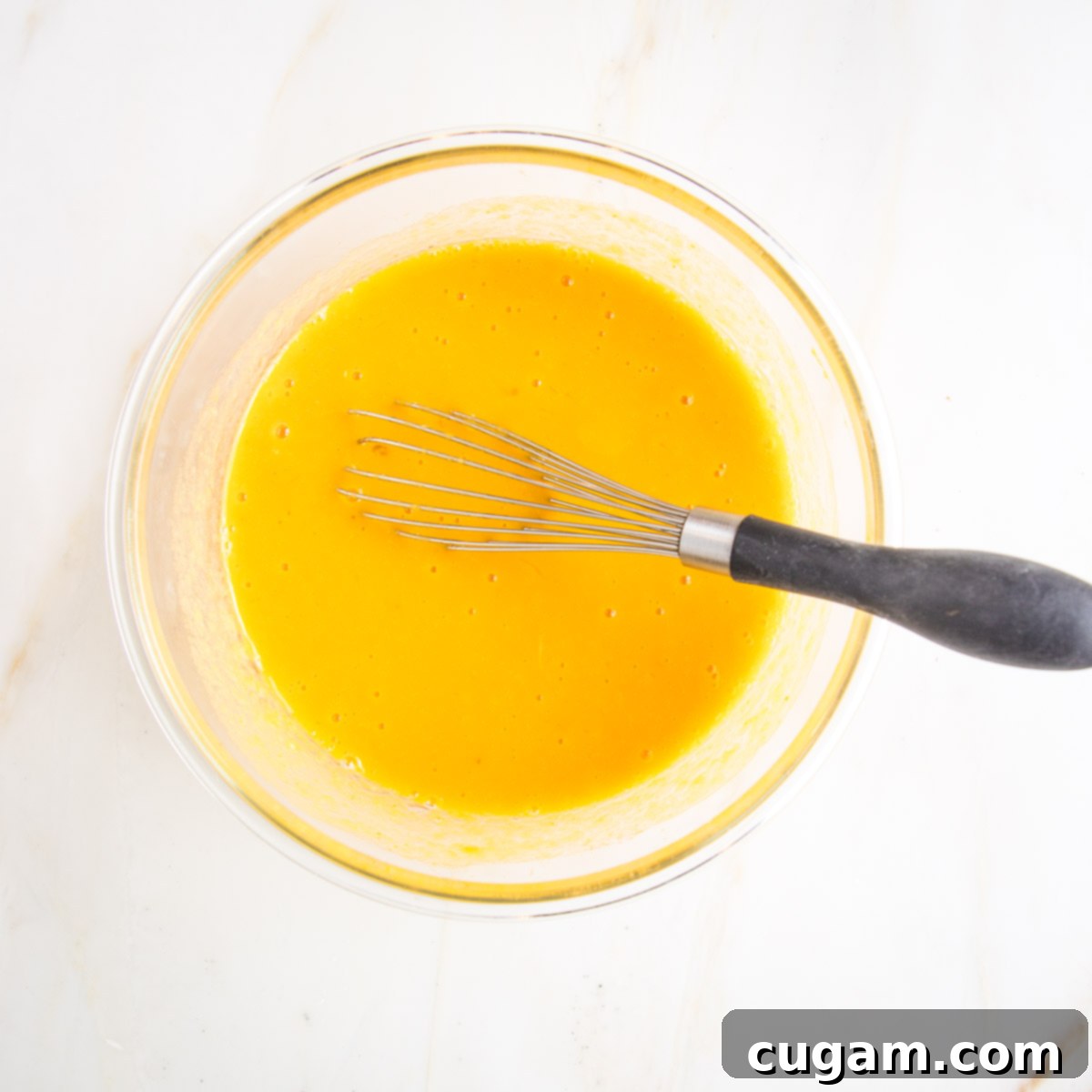A glass bowl filled with a yellowish liquid mixture, being whisked to combine the wet ingredients for the pumpkin cake.