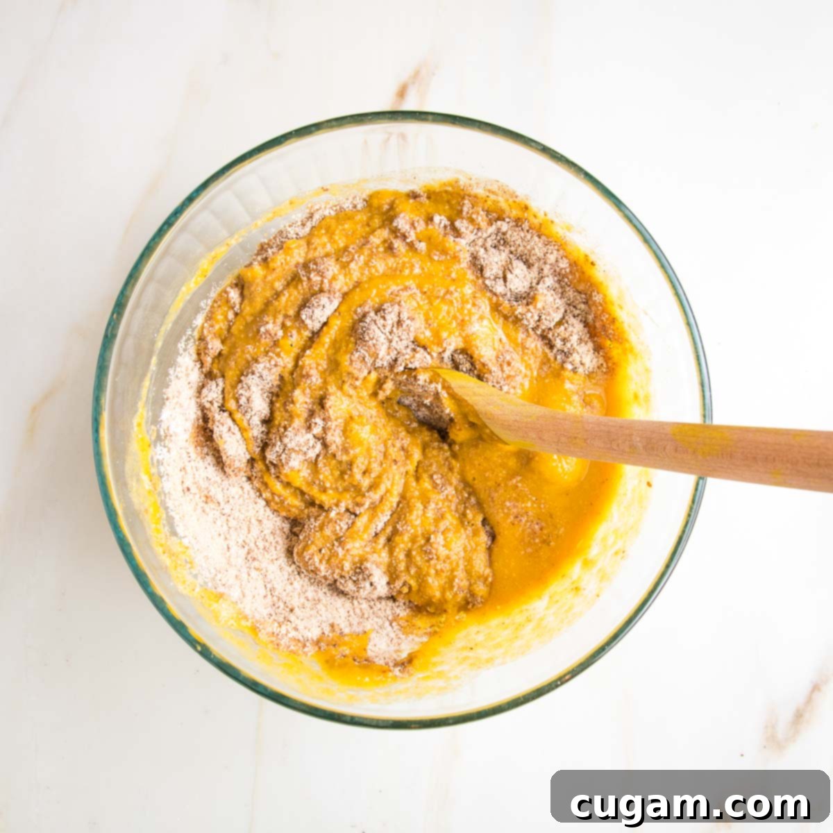 A glass bowl with a wooden spoon mixing together the wet and dry ingredients to form the pumpkin spice cake batter.
