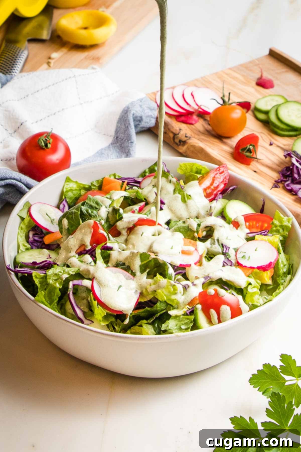 Simple salad with romaine, radishes, carrots, cucumbers and tomatoes in a white bowl. Vegan Ranch Dressing is being poured over the salad puddling in the middle.