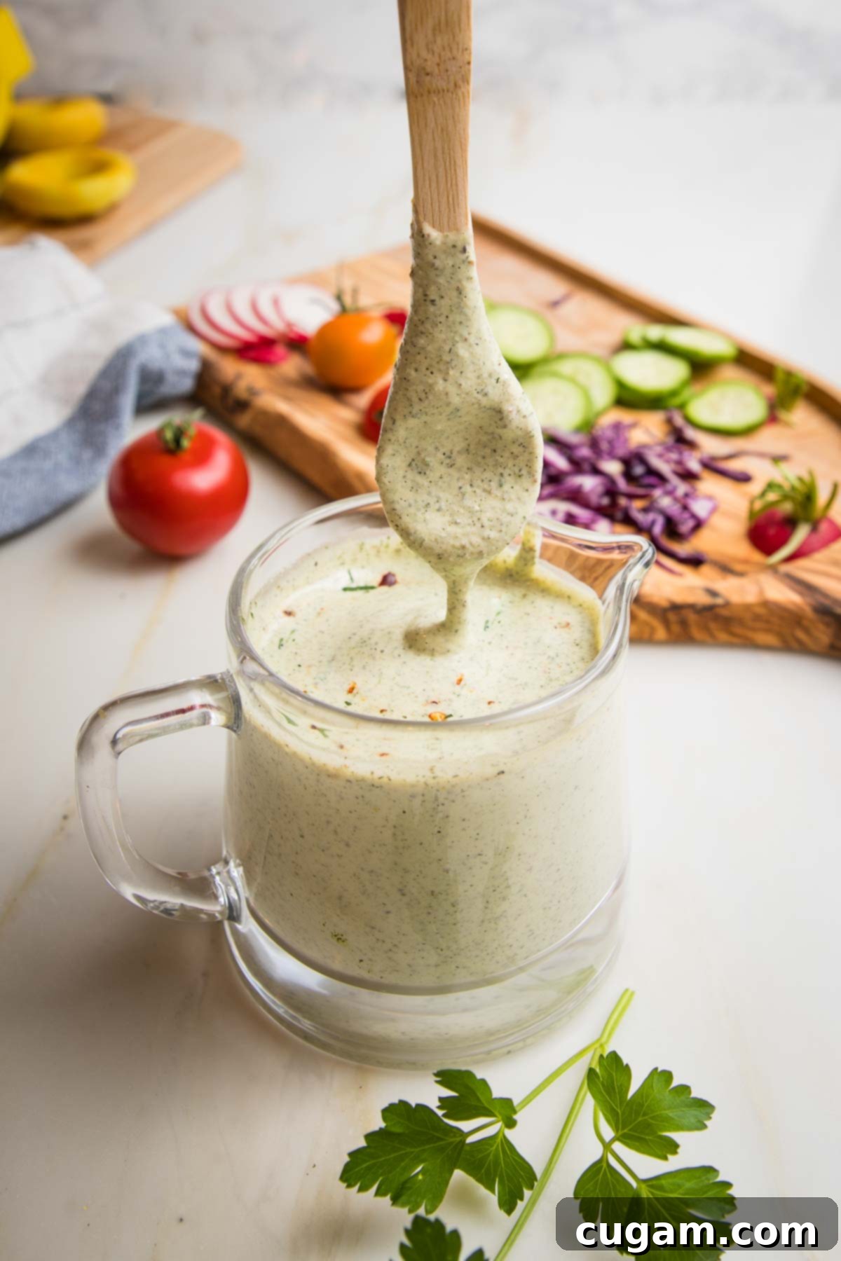Small glass pitcher filled with healthy ranch dressing. A wooden spoon is coming out of the dressing showing it to be drippy. There are random veggies on a cutting board in the background, ready for dipping.