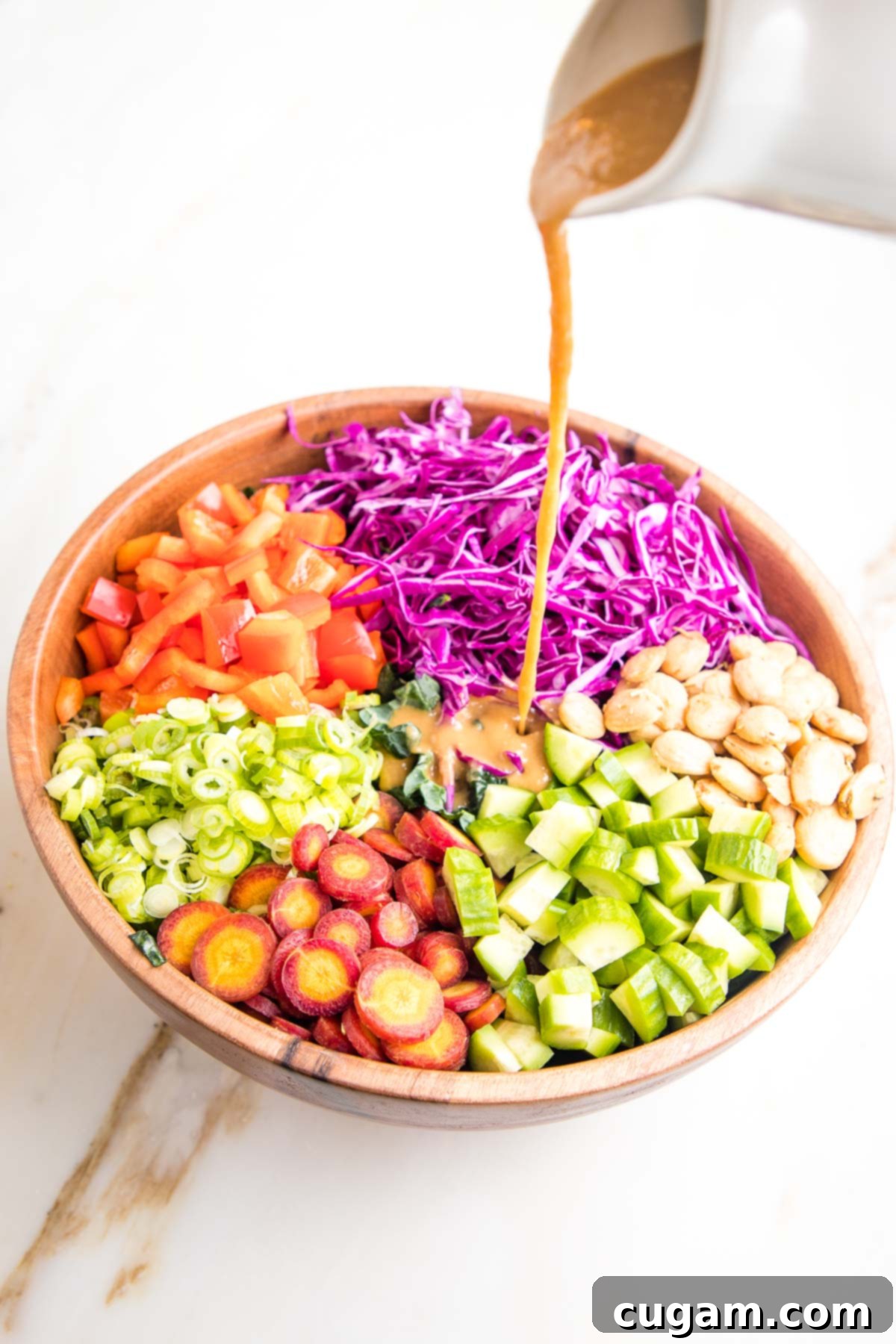 A wooden salad bowl featuring neatly arranged sections of colorful chopped vegetables (cabbage, cucumbers, carrots, red peppers, scallions, and Marcona almonds) over kale, with a small white pitcher pouring light brown dressing over the center.
