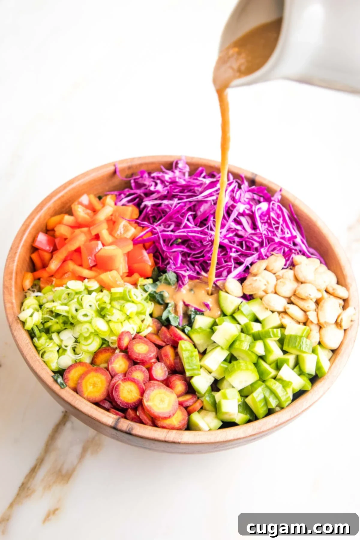 Wooden Salad bowl with sections of colorful chopped veggies cabbage, cucumbers, carrots, red peppers, scallions and marcona almonds) and a small white pitcher of light brown dressing pouring over the center of the bowl.
