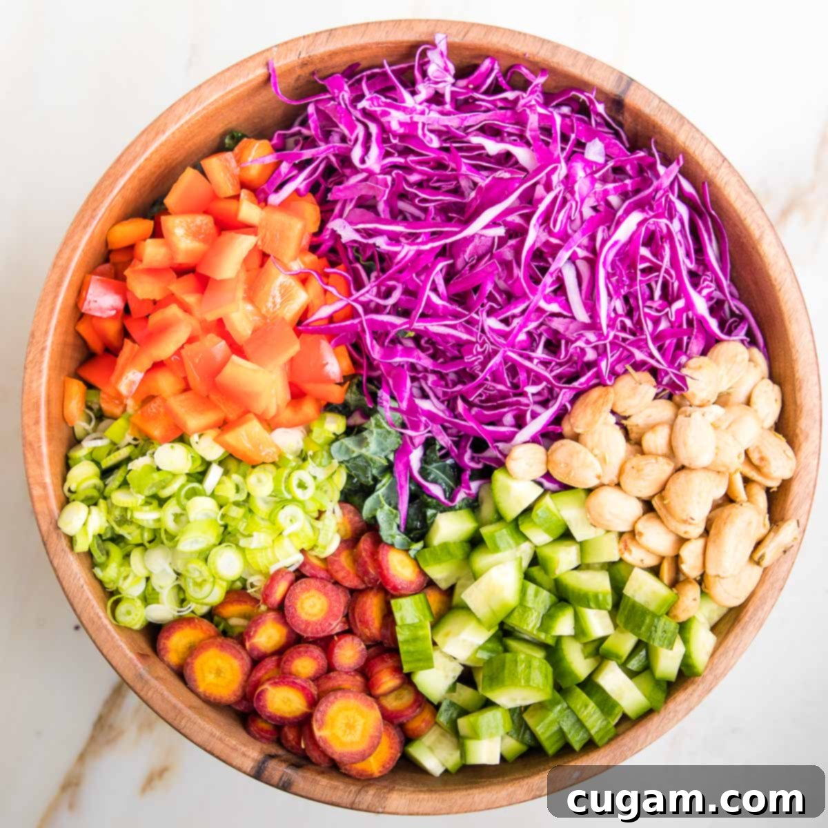 Wooden salad bowl filled with chopped kale and then sections of chopped veggies are on top of the kale. Purple cabbage, red peppers, scallions, purple carrots, cucumbers and marcona almonds