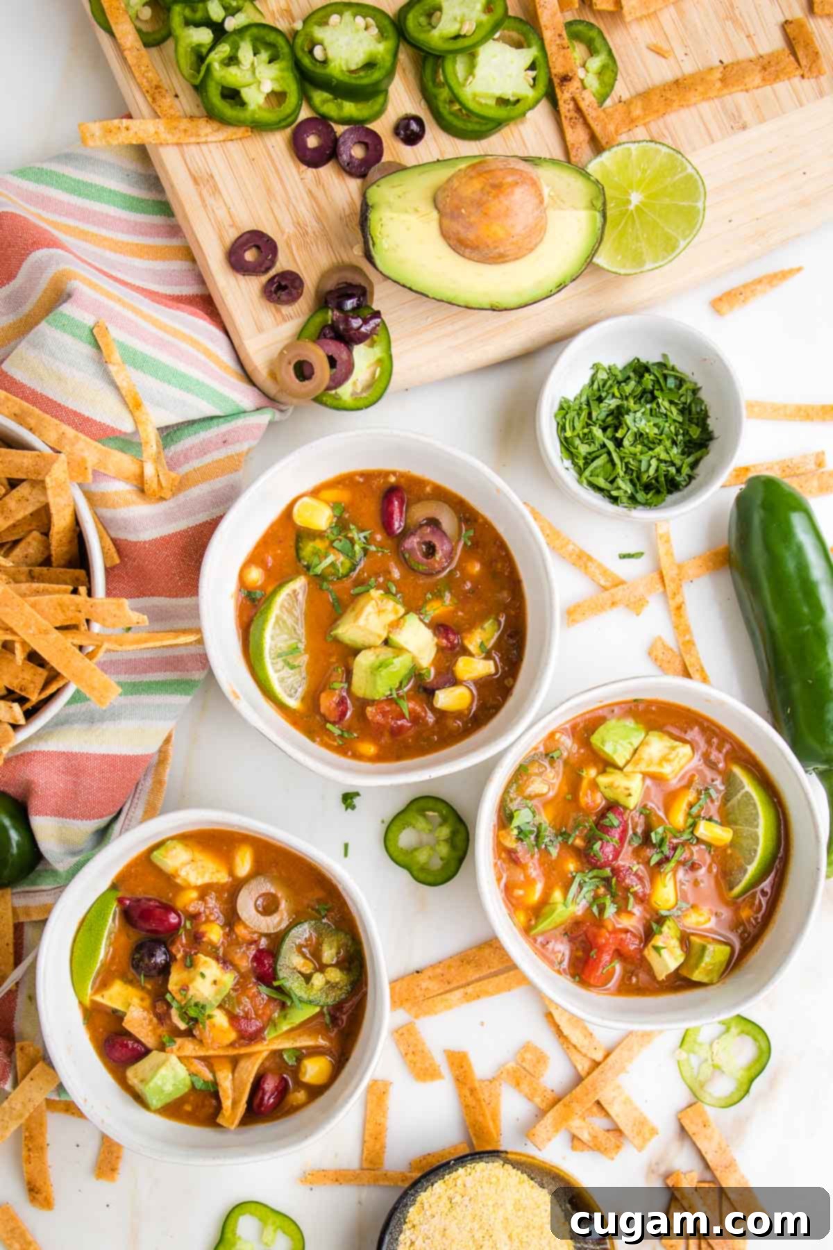 Three bowls of colorful vegan taco soup, with chunks of avocado, slices of jalapeno, some tortilla strips, a bowl of chopped cilantro, some nutritional yeast and the edge of a cutting board filled with sliced olives, jalapenos and an avocado. 