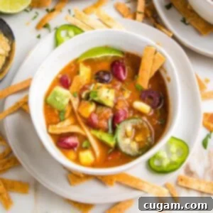 White bowl with plate underneath filled with vegan taco soup. It's colorful, with yellow corn, green jalapenos, chunks of avocado, red kidney beans and crispy tortilla strips. You also can see additional toppings strewn about on the white counter.