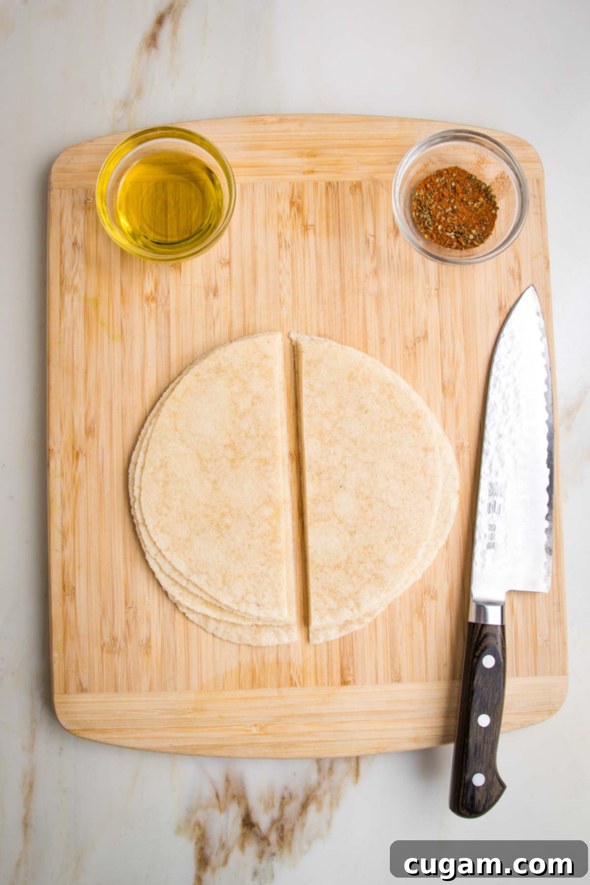 Wooden board with a small dish of olive oil and another small dish with taco seasonings. There are flour tortillas stacked in the center with a sharp knife off to the side, the tortillas have been cut in half.