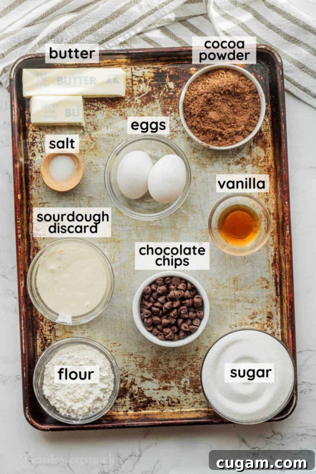 Carefully measured ingredients for sourdough discard brownies, including butter, cocoa, sugar, eggs, and sourdough discard, neatly arranged in glass bowls on a metal baking sheet.