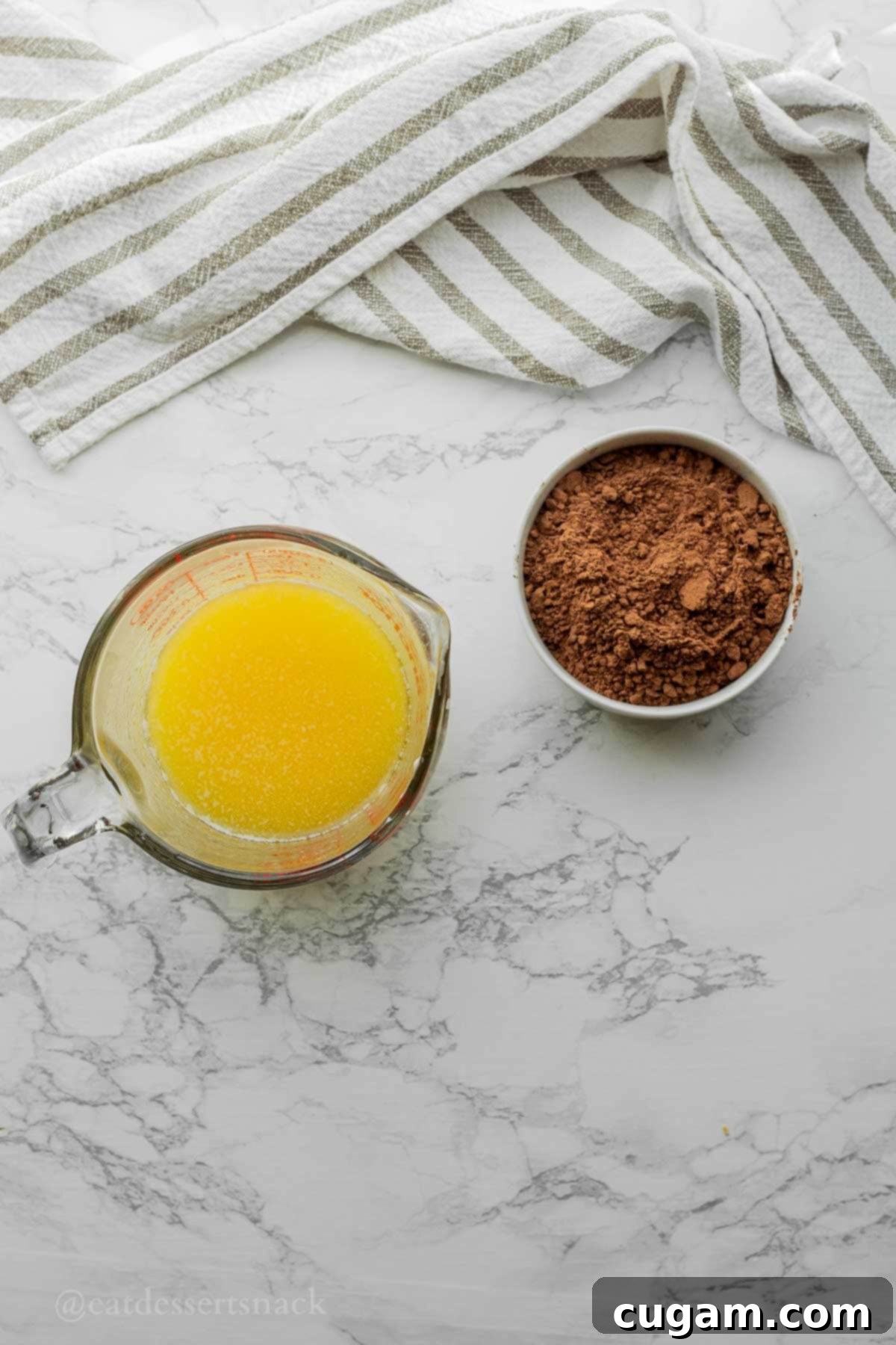 Melted butter in a glass measuring cup beside a white bowl of cocoa powder on a clean marble countertop, ready for blooming.