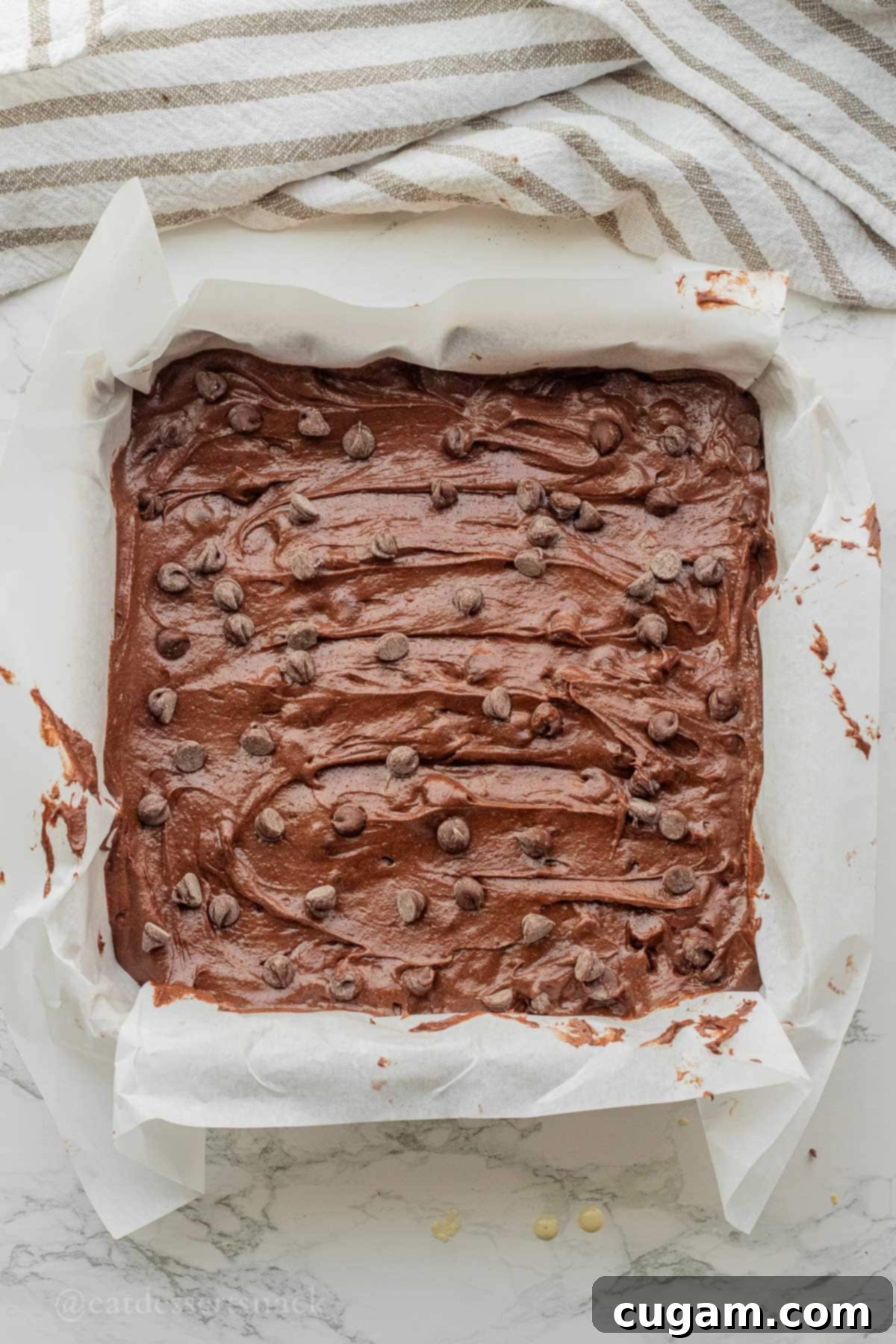 Raw sourdough discard brownie batter spread evenly in a parchment-lined 9x9 inch baking pan, generously sprinkled with semi-sweet chocolate chips on top, ready for baking.