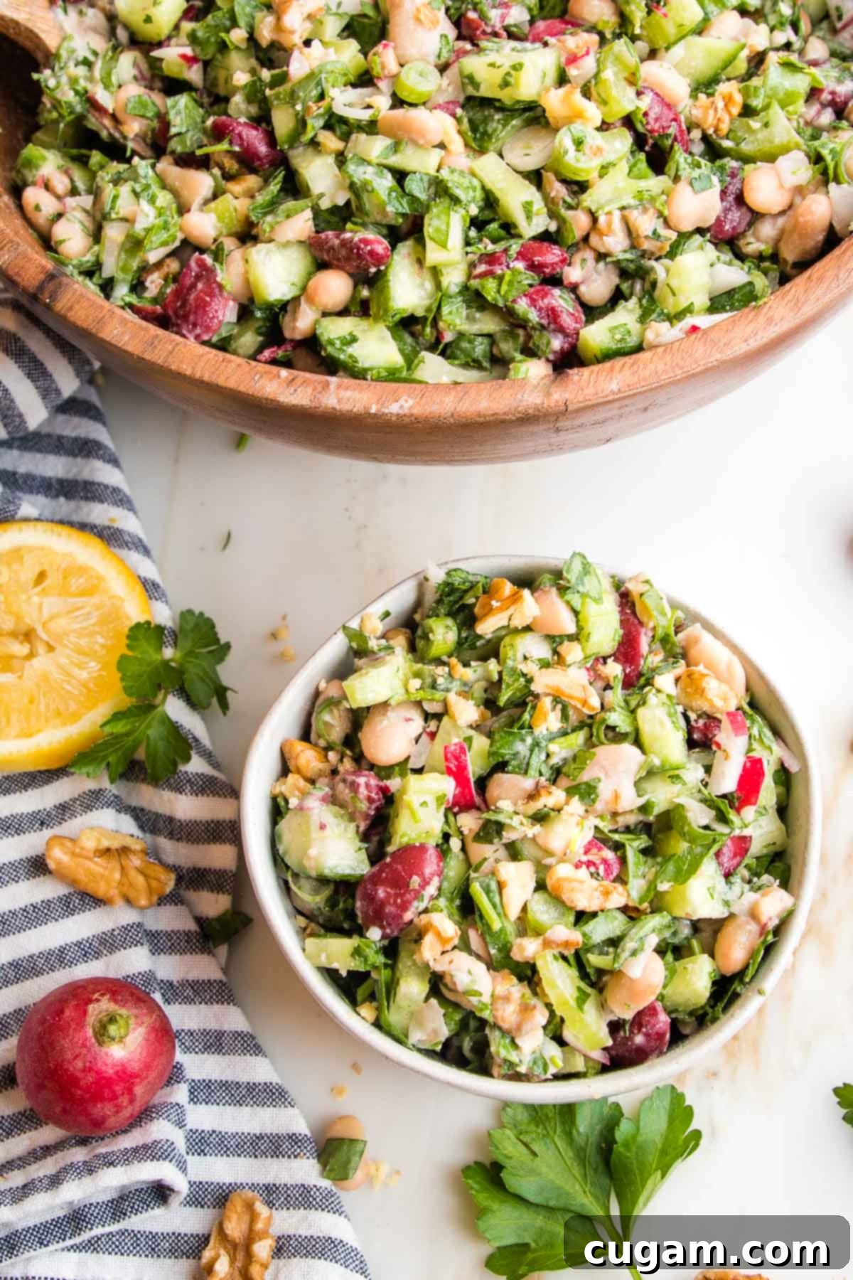Bean salad with greens and radishes.Large wooden bowl plus small white bowl with same salad. Whole lemon, walnuts, radish and fresh parsley leaves on the counter on a blue striped towel. 
