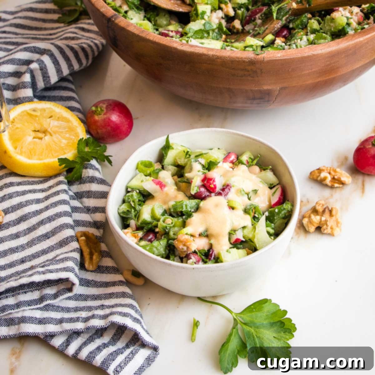 white bowl filled with bean salad with lemon tahini dressing.  larger bowl in the background along with lemon half, whole radishes and sprigs of fresh parsley.