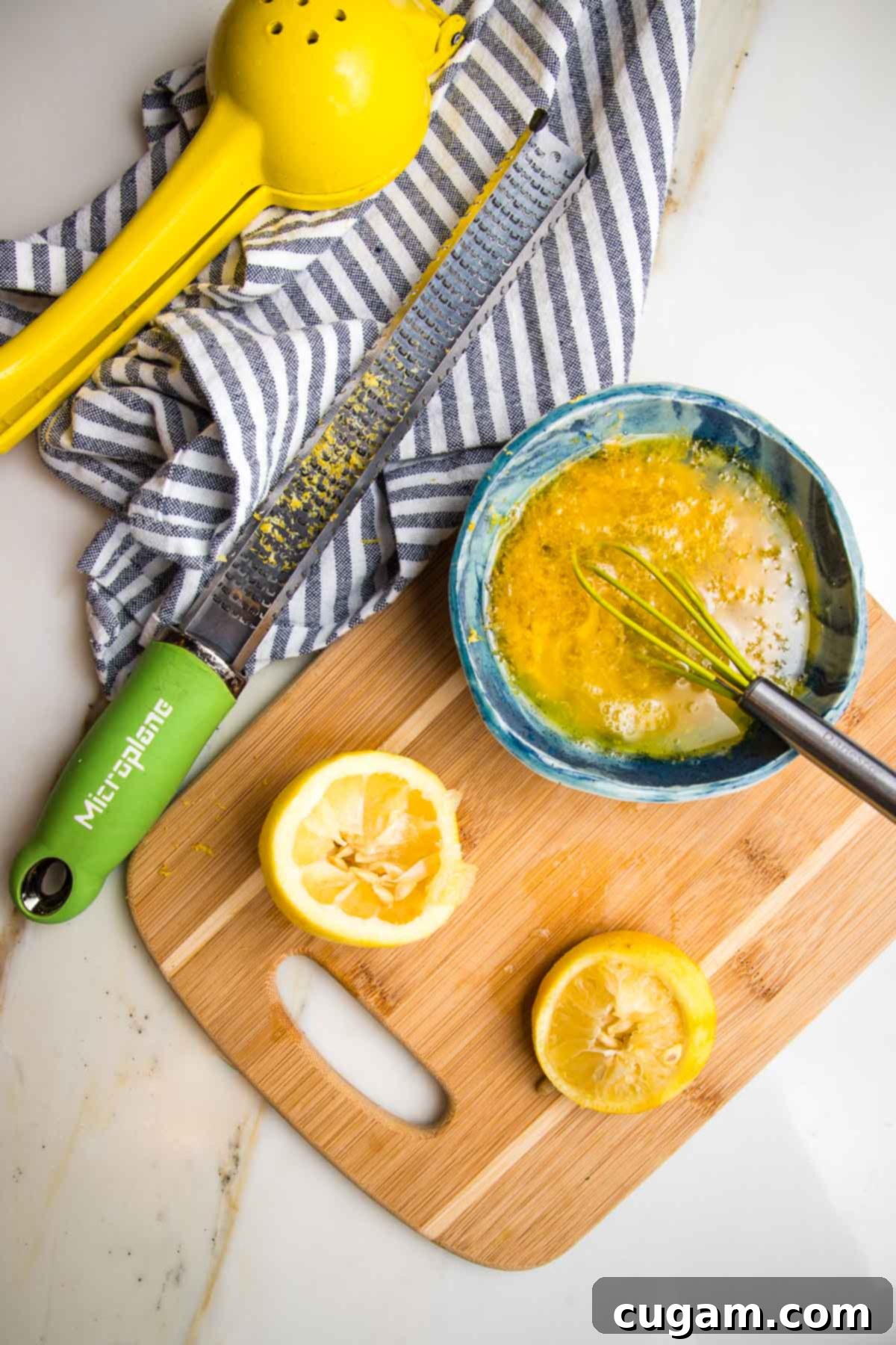 oily dressing whisked together in teal bowl. Two halves of a lemon already zested and squeezed lie on a cutting board. A zester with lemon zest on it on a blue striped towel.
