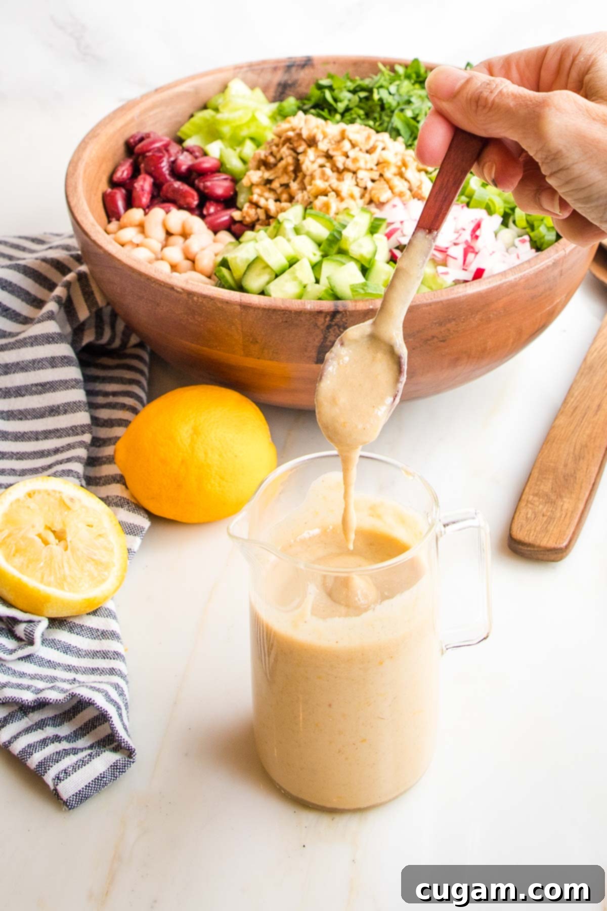 small glass pitcher with creamy lemony dressing. there are lemons in the distance along with a wooden bowl filled with chopped veggies.