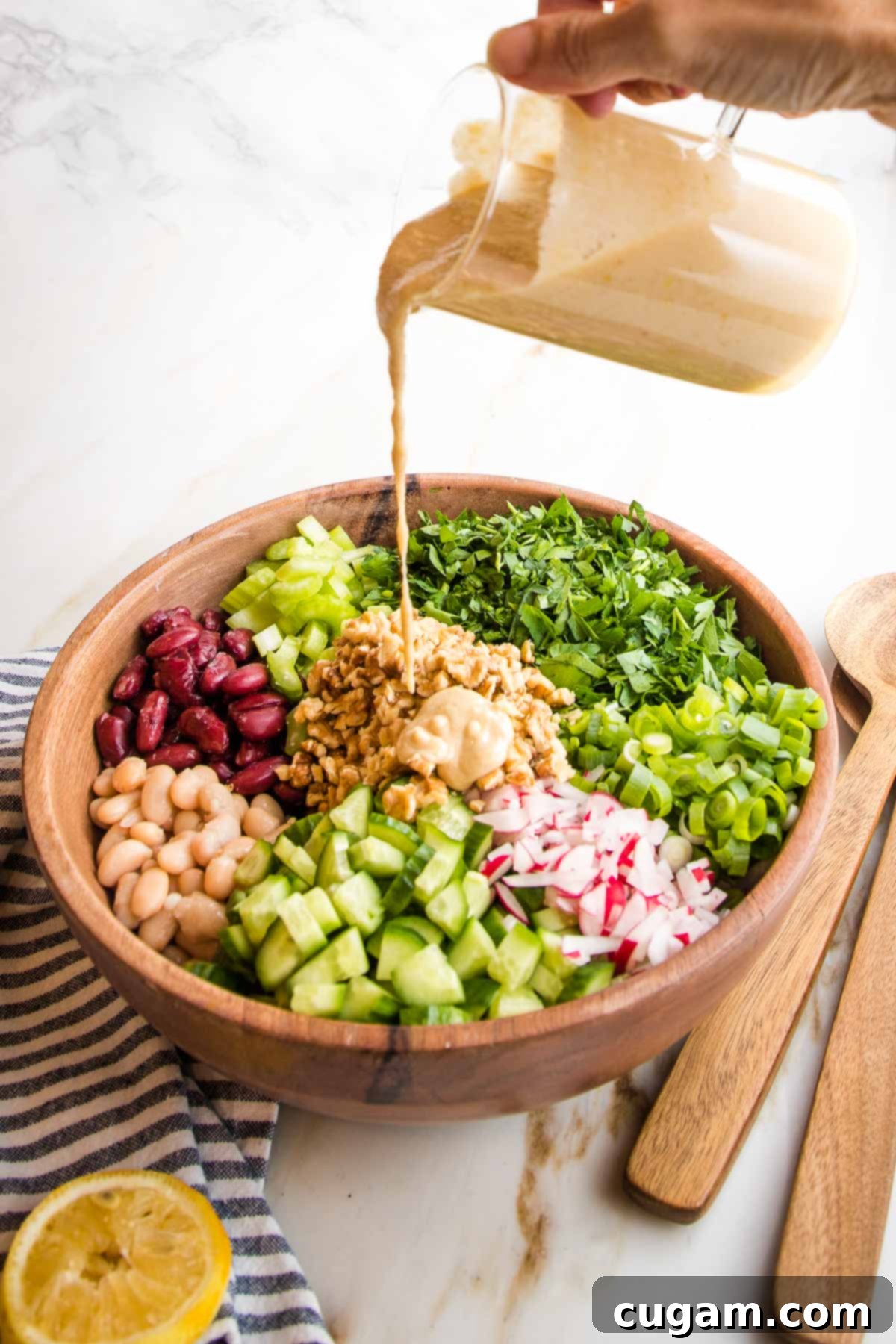Wooden bowl with vibrant chopped vegetable sections. Lemon tahini dressing is being poured over the bowl at the center where the walnuts are.