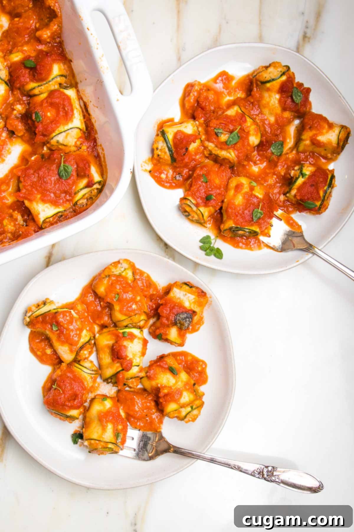 White plate with zucchini roll ups filled with ricotta and topped with marinara. Another plate full and the corner of the baking dish can be seen.
