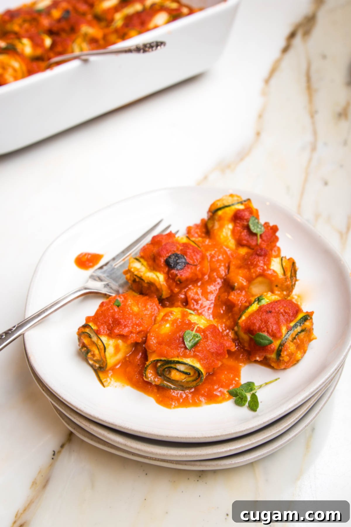 White rimmed plate filled with rolled up zucchini lasagna rolls covered in marinara sauce. Fork on the plate. Casserole dish filled with more visible in the background.