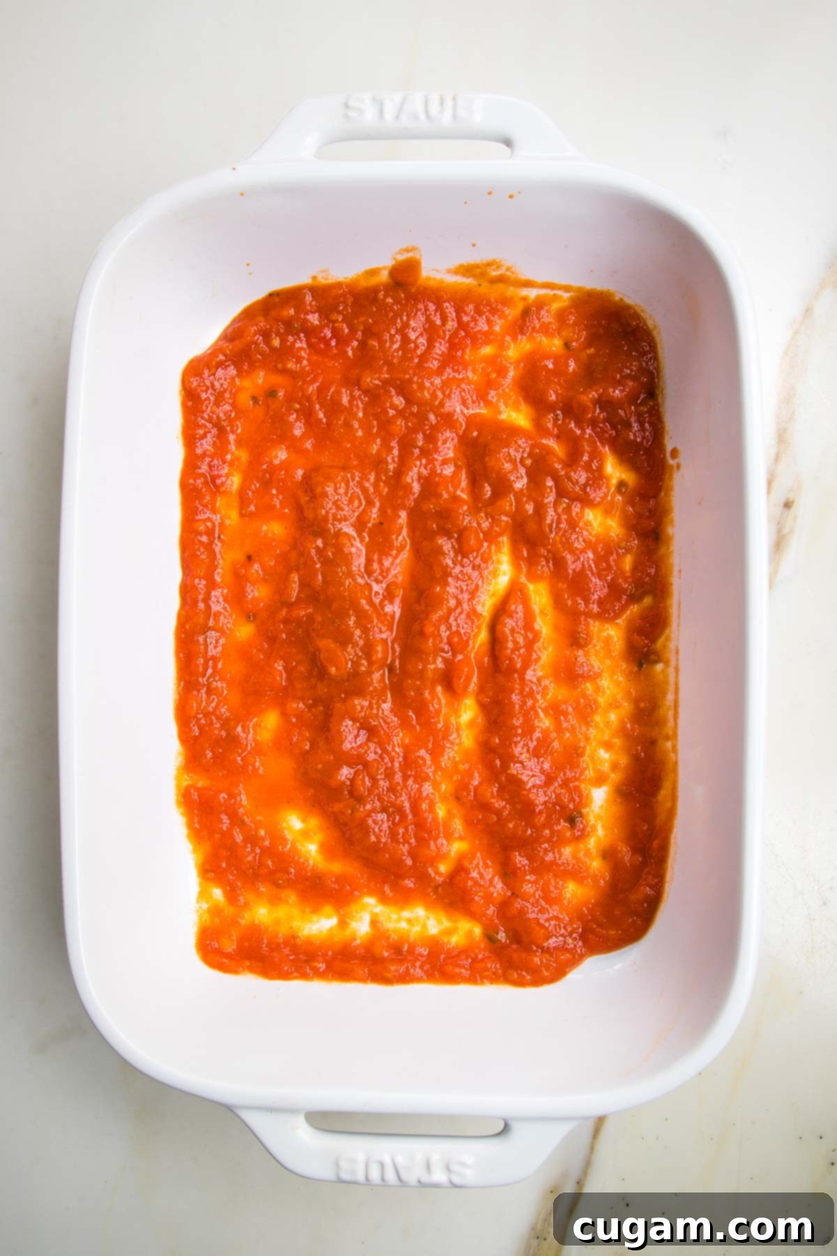 White baking pan with a thin layer of bright red marinara sauce spread on the bottom