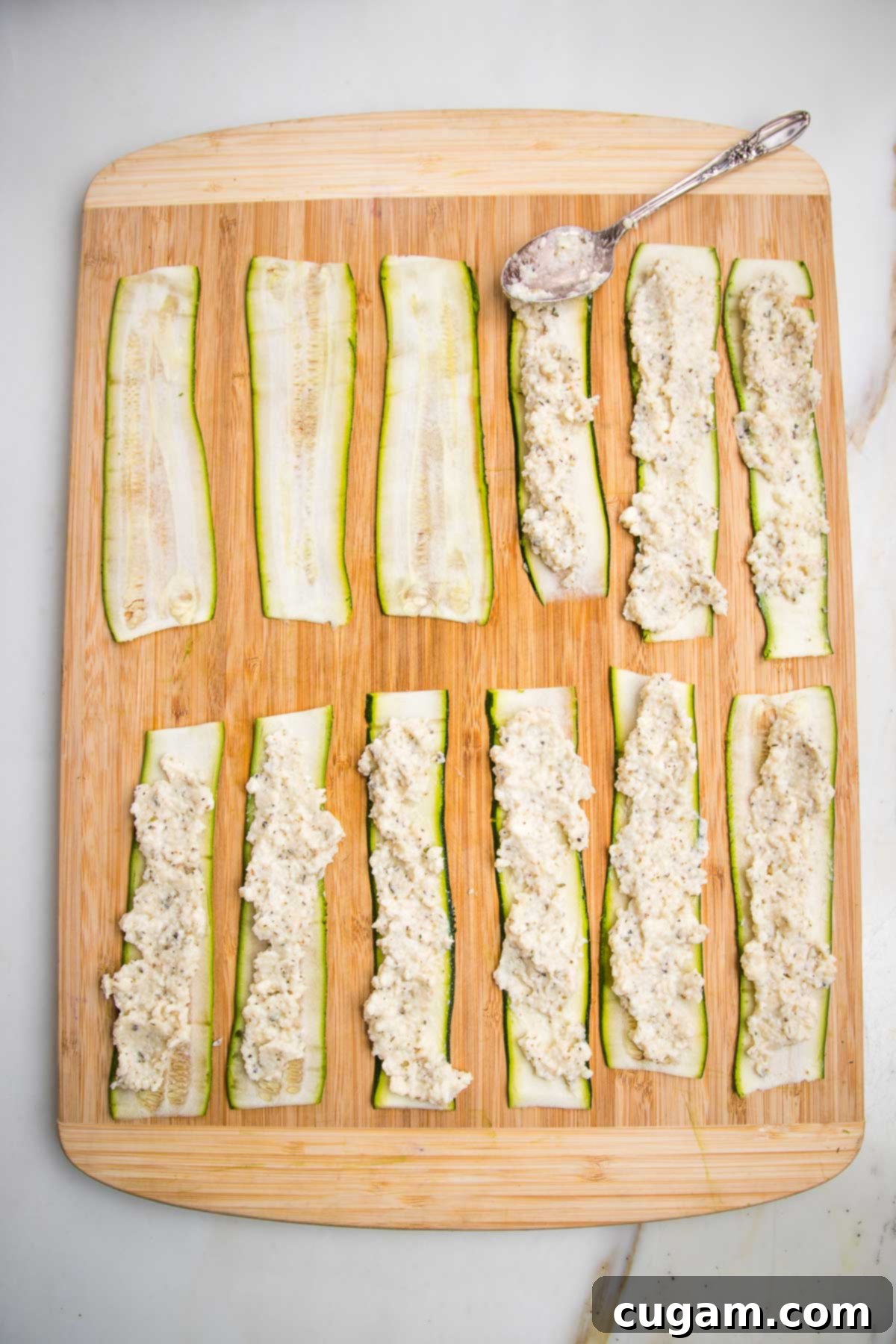 Wooden board with zucchini strips laid out, a teaspoon full of ricotta, spread onto each strip is in process.