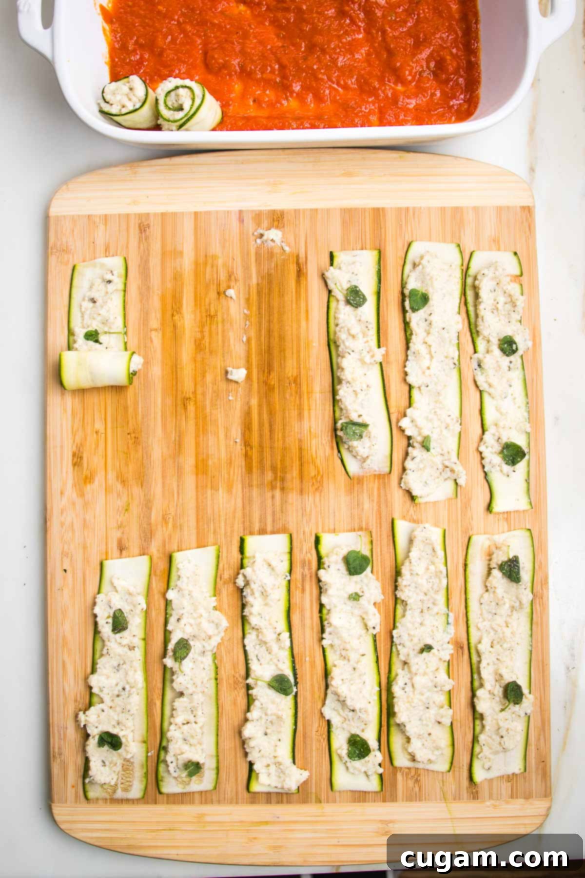 Cutting board with zucchini strips spread with ricotta, one is shown to be in process of rolled up and you can see a baking pan with marinara sauce where the finished rolls are being placed.