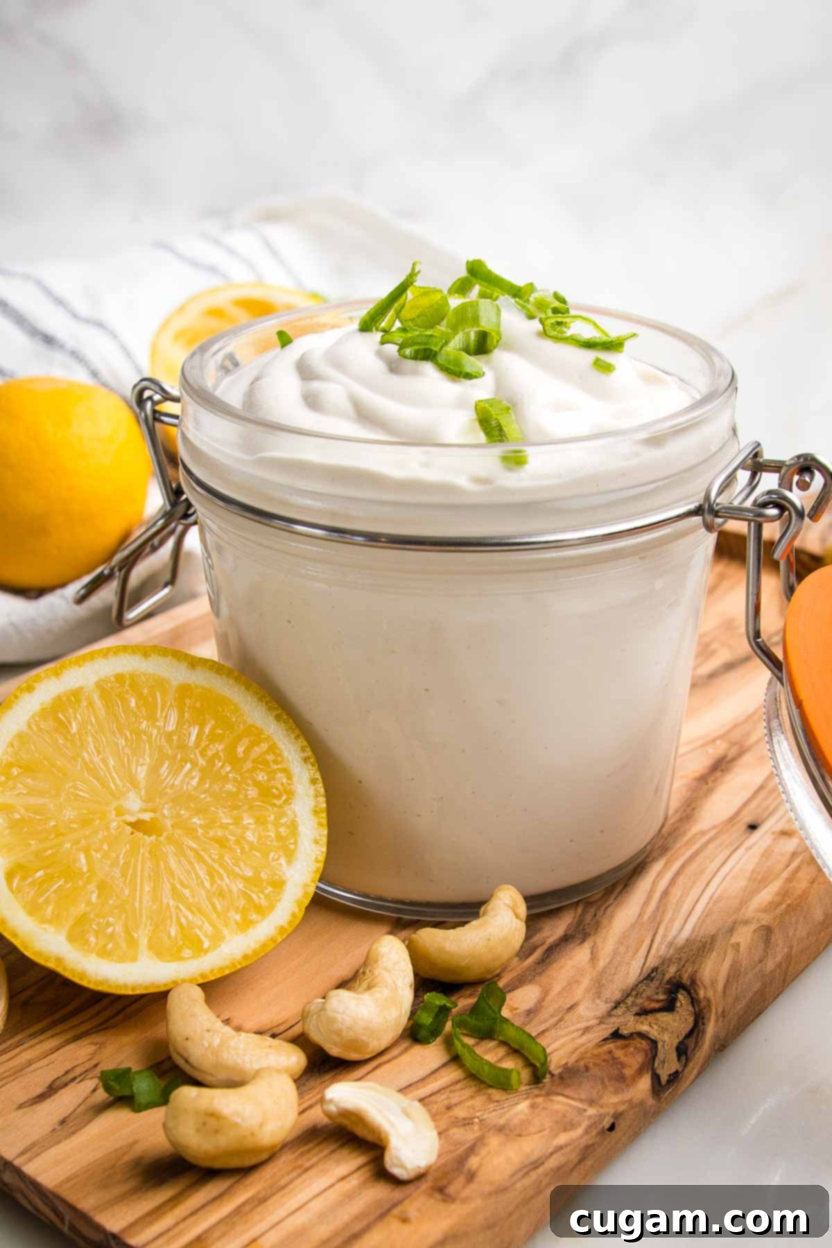 Vegan sour cream loaded into a glass canister and topped with scallions. All is on a wooden board with fresh lemon half and raw cashews. 