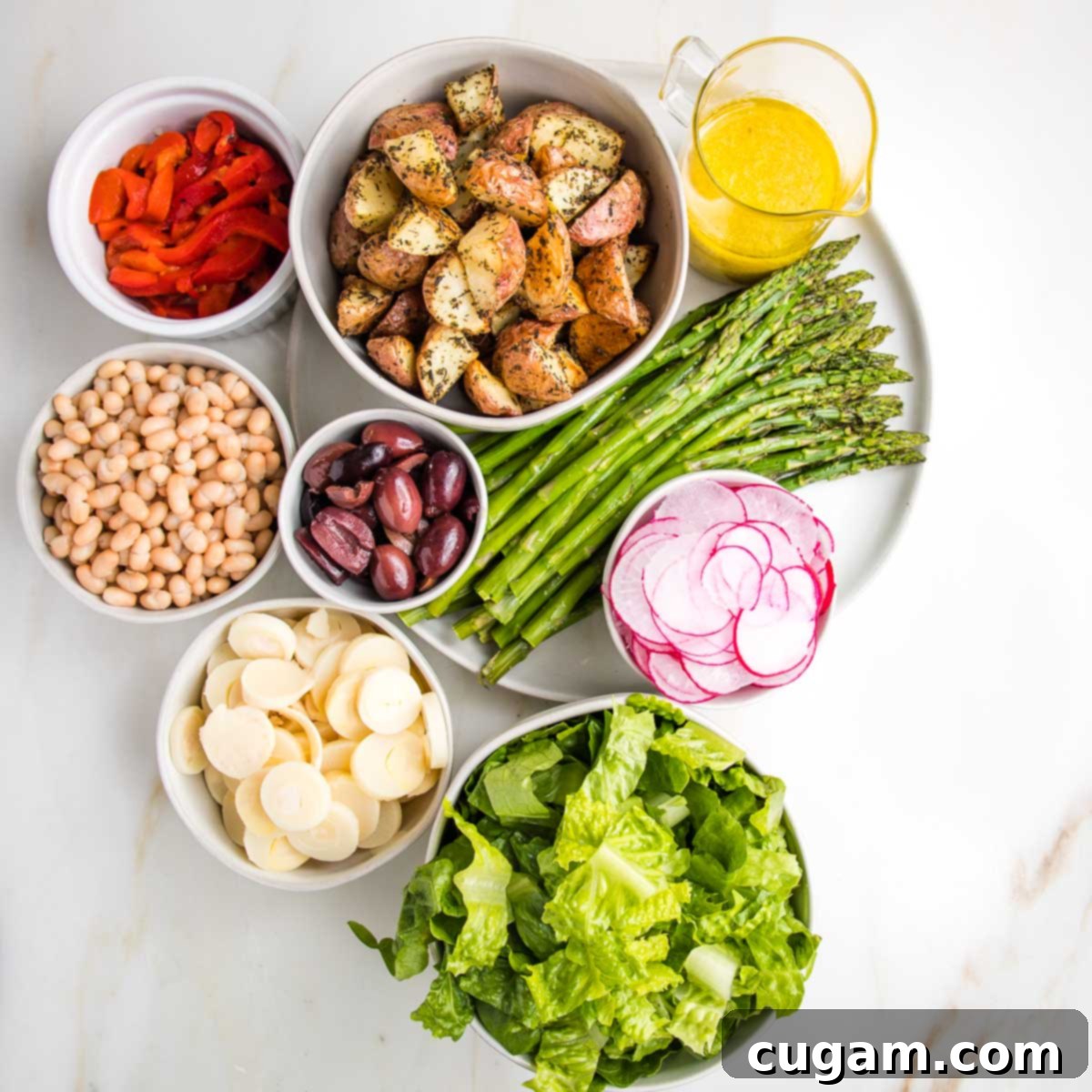 White dishes with separate ingredients for nicoise salad: roasted red pepper strips, roasted potatoes, lemon vinaigrette, roasted asparagus, small white beans, kalamata olives, slice hearts of palm, sliced radish and romaine lettuce.