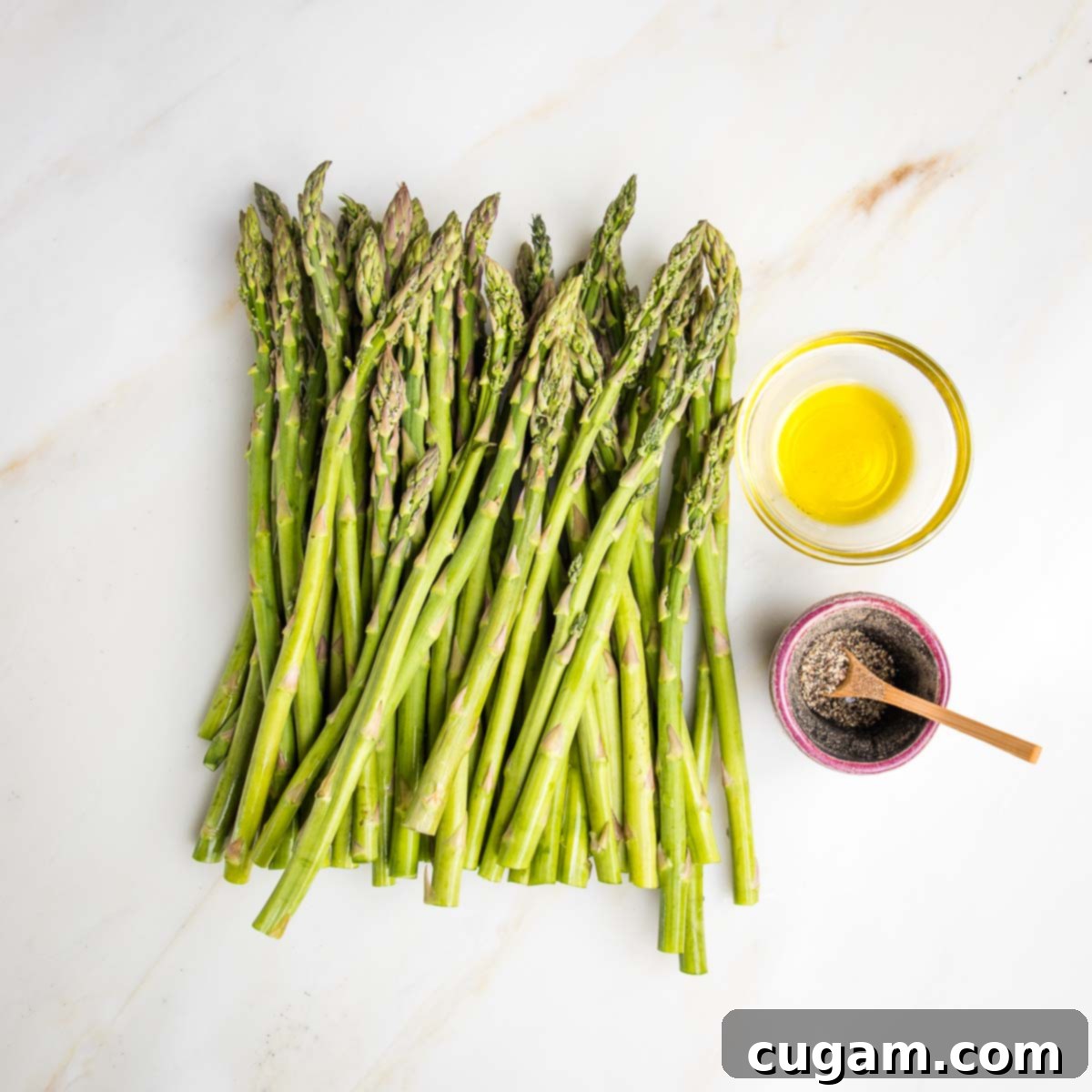 Fresh asparagus and a small glass dish of olive oil and a little container with fresh black pepper and a small wooden spoon.