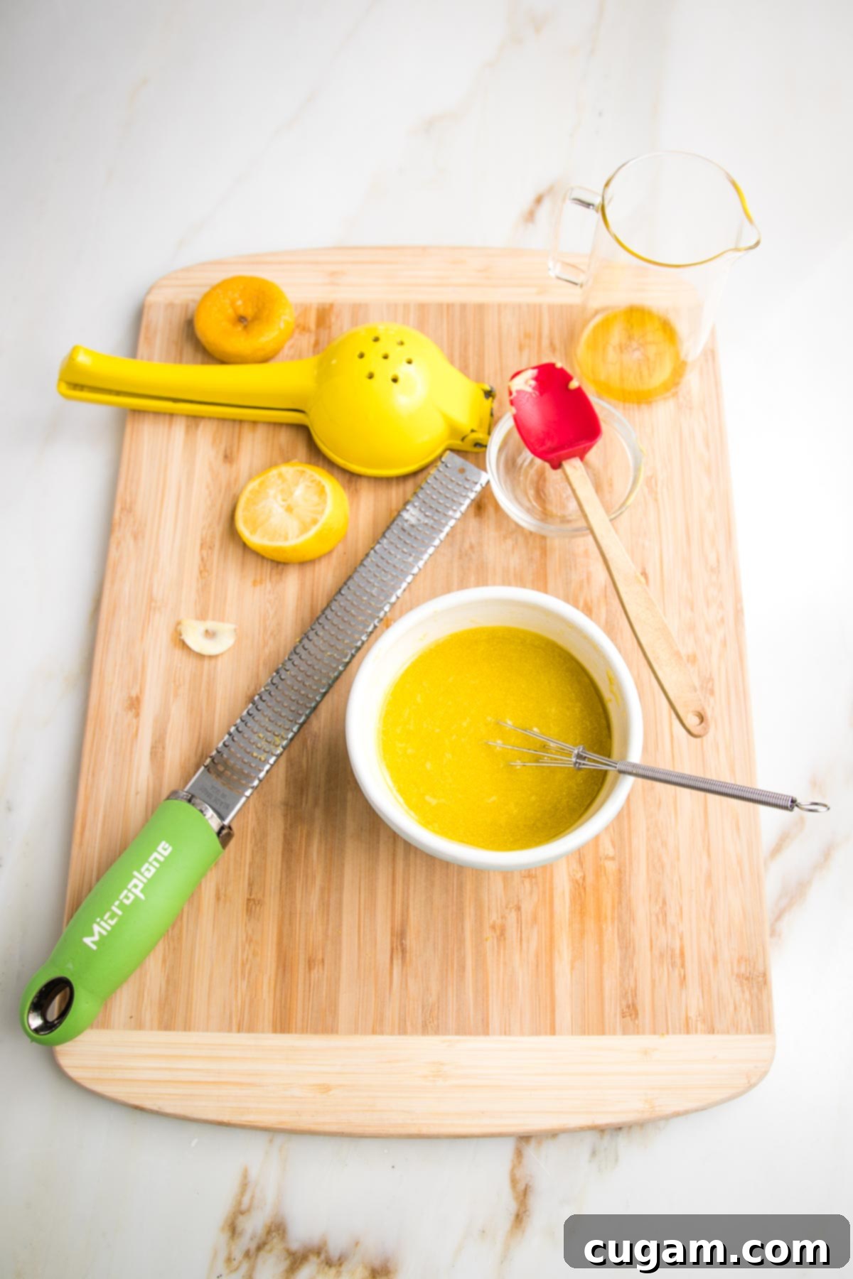 Lemon Vinaigrette made in a small bowl with a whisk. Also on the cutting board is a lemon squeezer, zester, lemon halves, spent garlic clove and pitcher of oil.