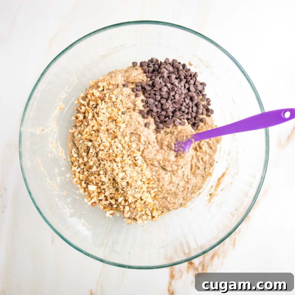 Vegan Zucchini Muffins 11 Zucchini muffin batter in a glass mixing bowl with a purple spatula and mini chocolate chips plus walnuts on top, ready to be folded in.