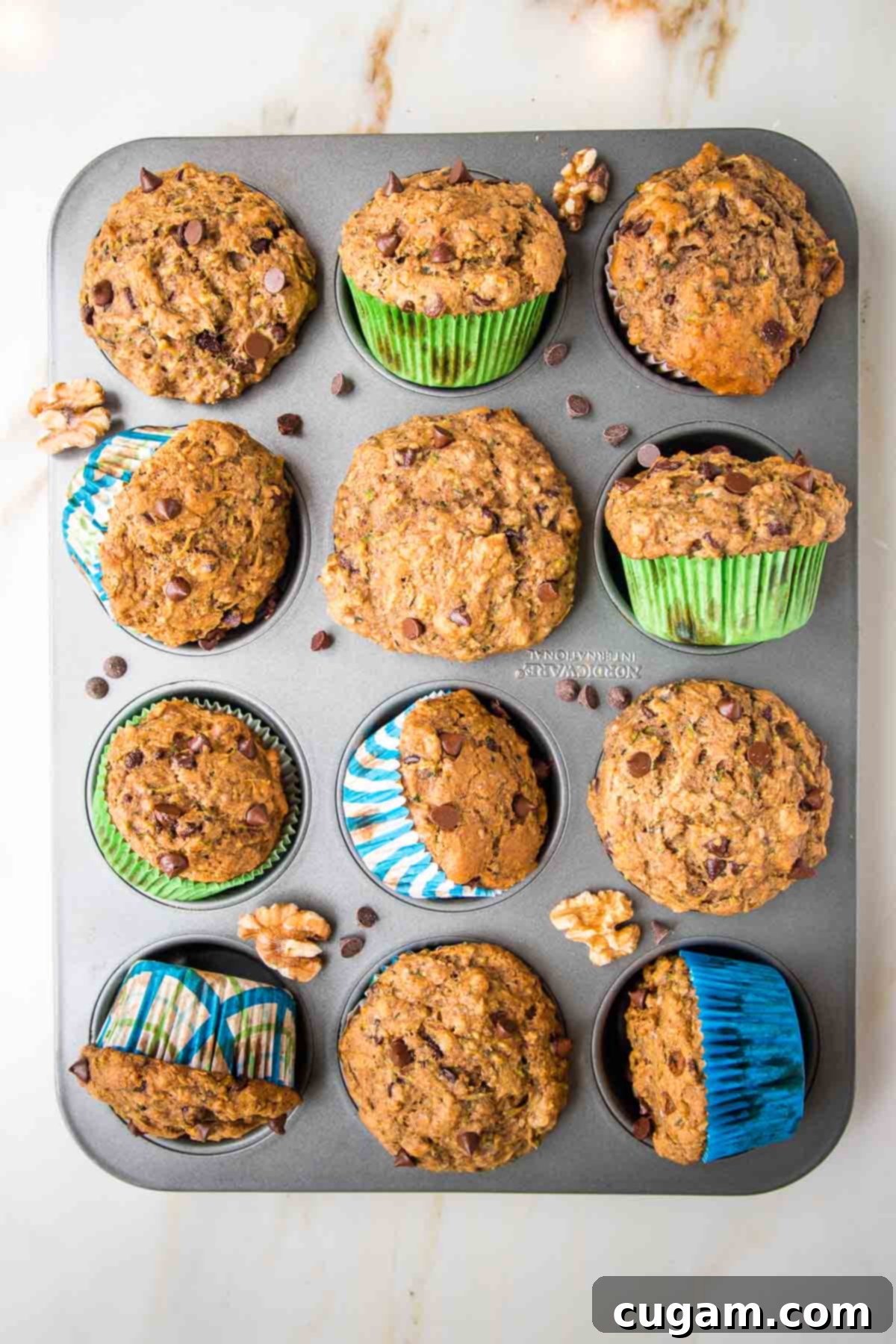 Vegan Zucchini Muffins 14 Chocolate Chip Zucchini Muffins fresh out of the oven in a muffin tin. Some are turned on their side and there are mini chocolate chips and walnuts strewn about, showcasing their golden-brown tops.