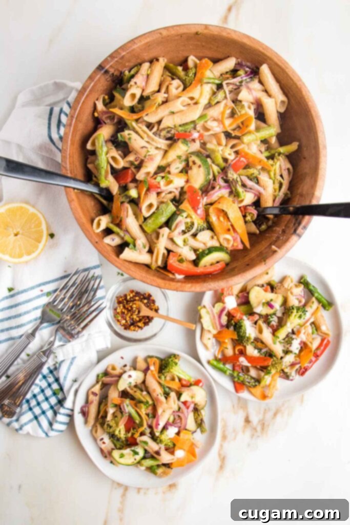 Brightly colored veggies plus penne pasta in a large wooden bowl with tossers.  There are two small plates served with same vegan pasta primavera, some forks, crushed red pepper and a half a lemon. 