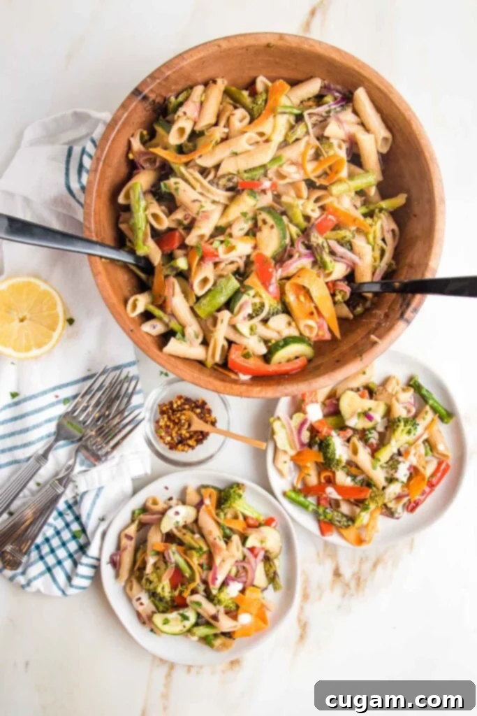 Brightly colored veggies plus penne pasta in a large wooden bowl with tossers.  There are two small plates served with same vegan pasta primavera, some forks, crushed red pepper and a half a lemon. 