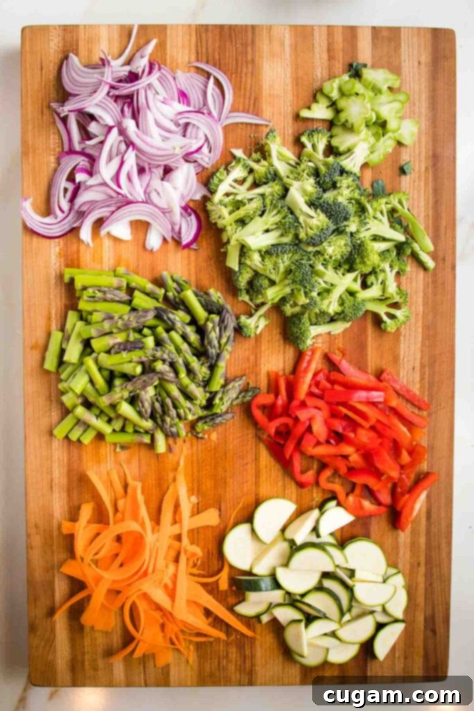 Wooden board with sliced red onion, broccoli florets, asparagus, red bell pepper, carrots and zucchini half moons. 