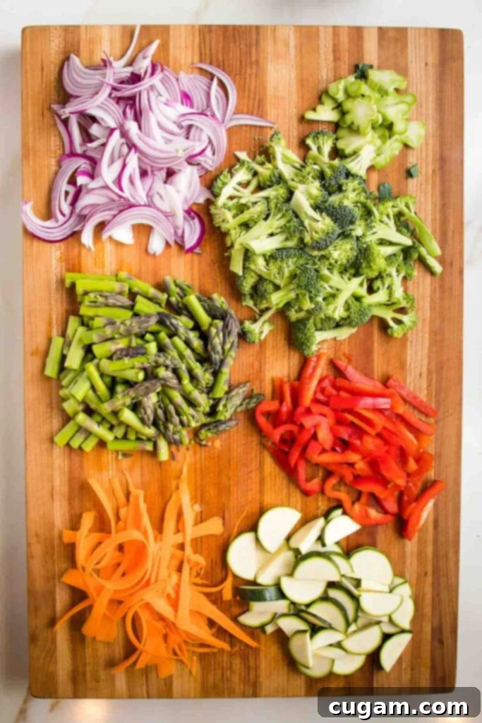 Wooden board with sliced red onion, broccoli florets, asparagus, red bell pepper, carrots and zucchini half moons. 