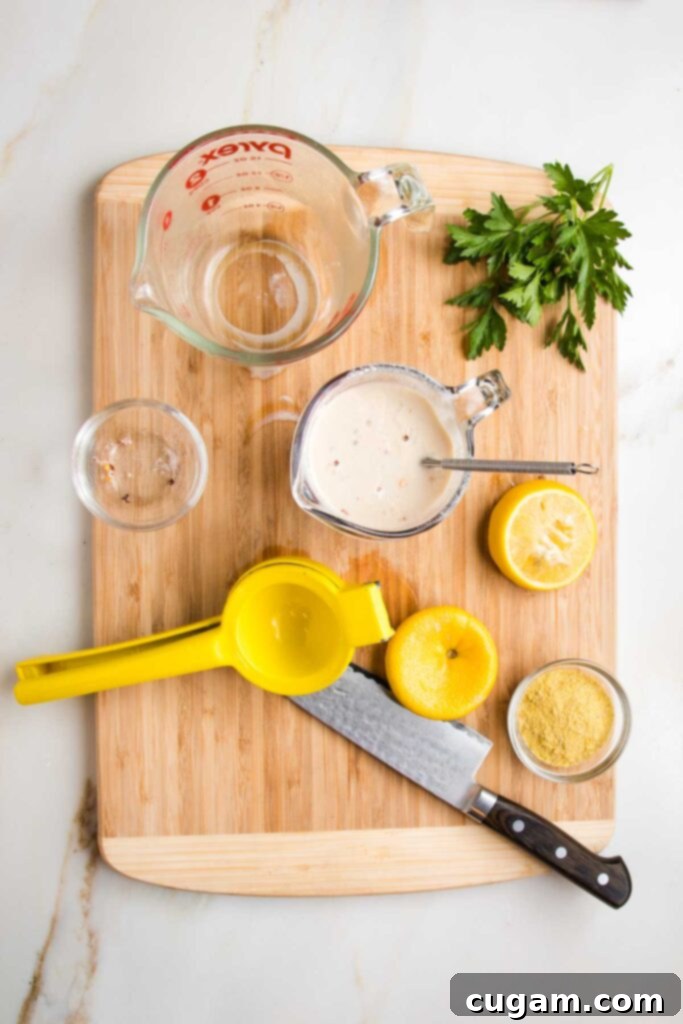 Wooden board with various glass dishes, a lemon squeezer, sharp knife, fresh parsley bunch, used up lemon halves, nutritional yeast and a pyrex measuring cup with a whisk, making a white sauce.