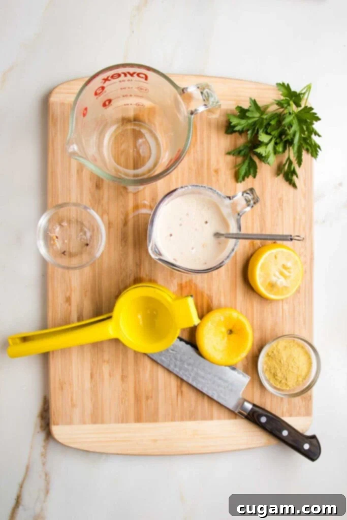 Wooden board with various glass dishes, a lemon squeezer, sharp knife, fresh parsley bunch, used up lemon halves, nutritional yeast and a pyrex measuring cup with a whisk, making a white sauce.