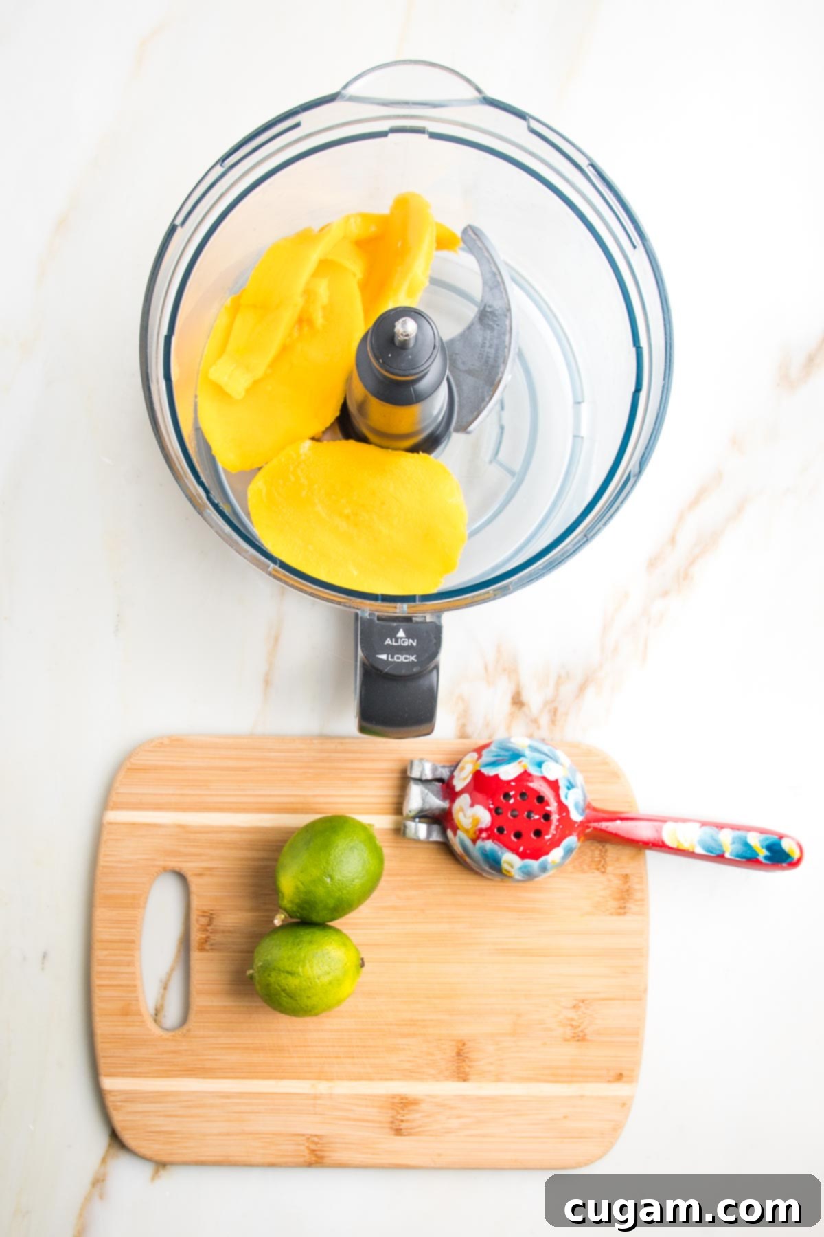 Food processor bowl with chunks of mango. Small wooden board with two limes and a colorful juice squeezer. 