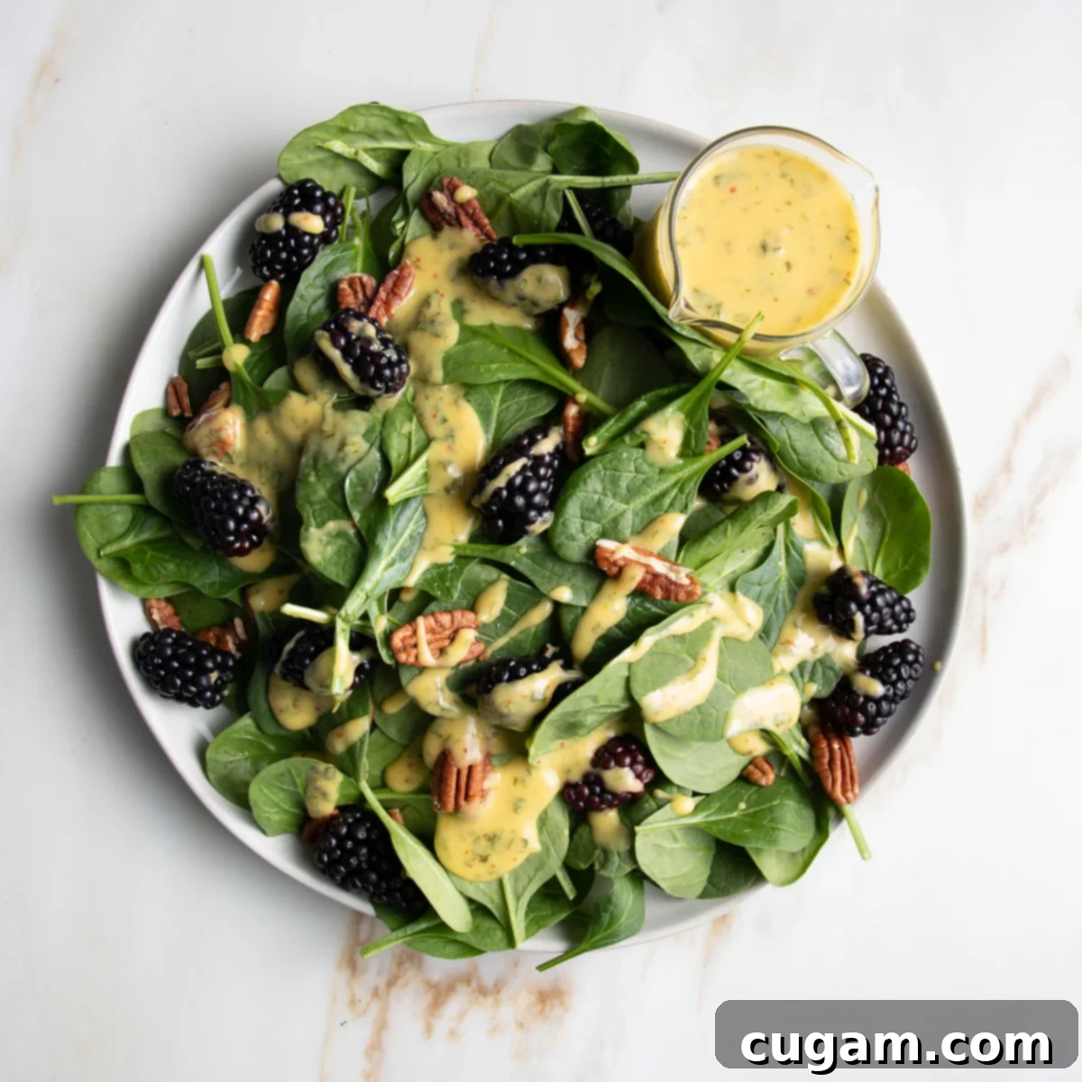 Spinach salad with blackberries, pecans and drizzled with mango dressing.  There is a small glass pitcher with extra yellow, mango dressing on the side of the round white rimmed plate. 