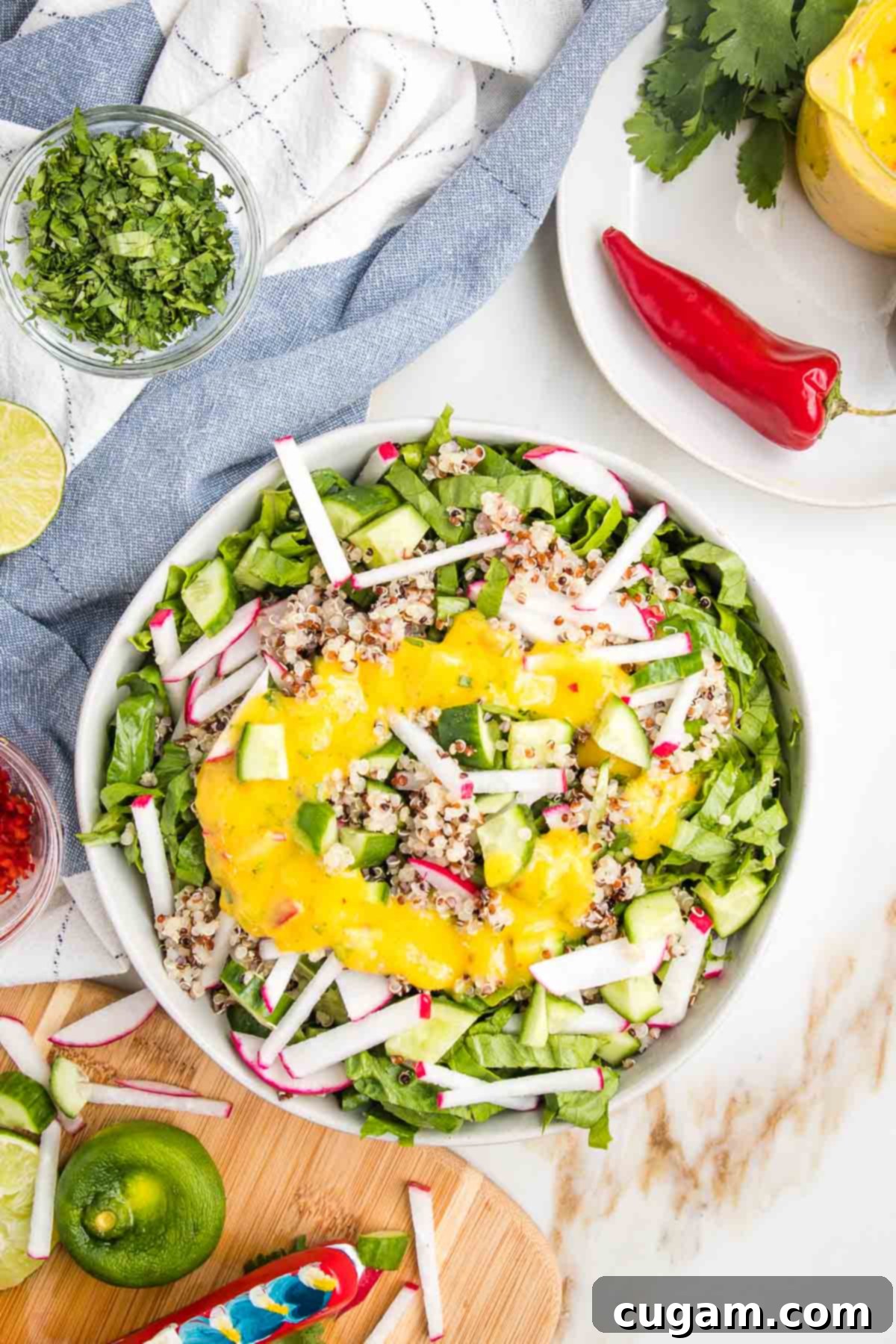 Large white salad bowl filled with julienned romaine, radishes, cucumbers, drizzled with bright yellow mango salad dressing. Dispersed on the counter are cilantro, limes, red hot chili peppers and chunks of veggies.
