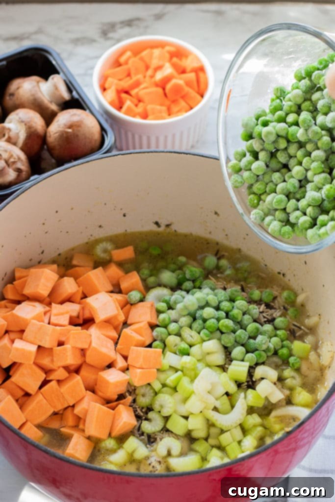 A cook's hands pouring a bowl of diced butternut squash, carrots, peas, and mushrooms into a large pot containing sautéed chicken, onions, and broth with wild rice.