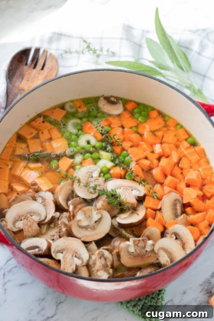 A large pot on the stovetop filled with unmixed ingredients for creamy chicken wild rice soup, including raw chicken, sliced mushrooms, peas, diced carrots, squash, wild rice, and fresh herbs.