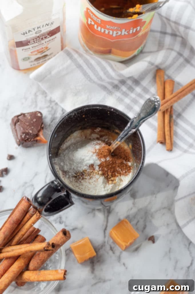 Ingredients for a pumpkin mug cake laid out next to a 12 oz. mug. Components include milk, pumpkin puree, vanilla, flour, sugar, and various fall spices.