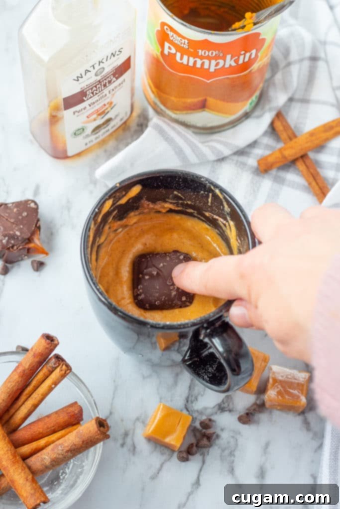 Hand pushing a chocolate-covered caramel into raw pumpkin mug cake batter in a mug.